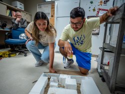 Two students kneel behind a small square maze. Their professor sits behind them.