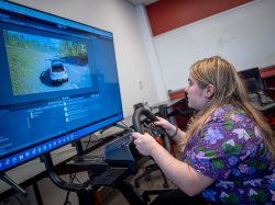 Student seated at a steering wheel looks at a computer screen with an image of a car on a highway.