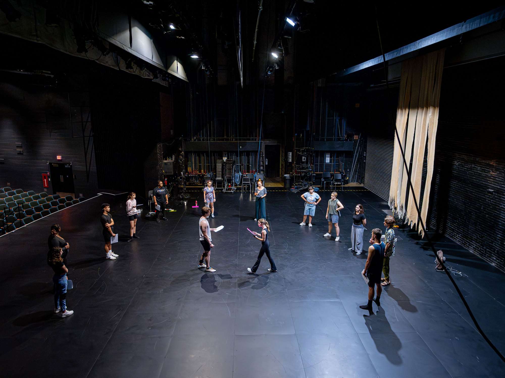 A group of 10 actors form a semi-circle on a black stage while two actors read from scripts in the middle of the stage.