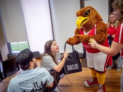 A college mascot, a giant red hawk, gives a student a black bag with the words “Day of Service” on it.