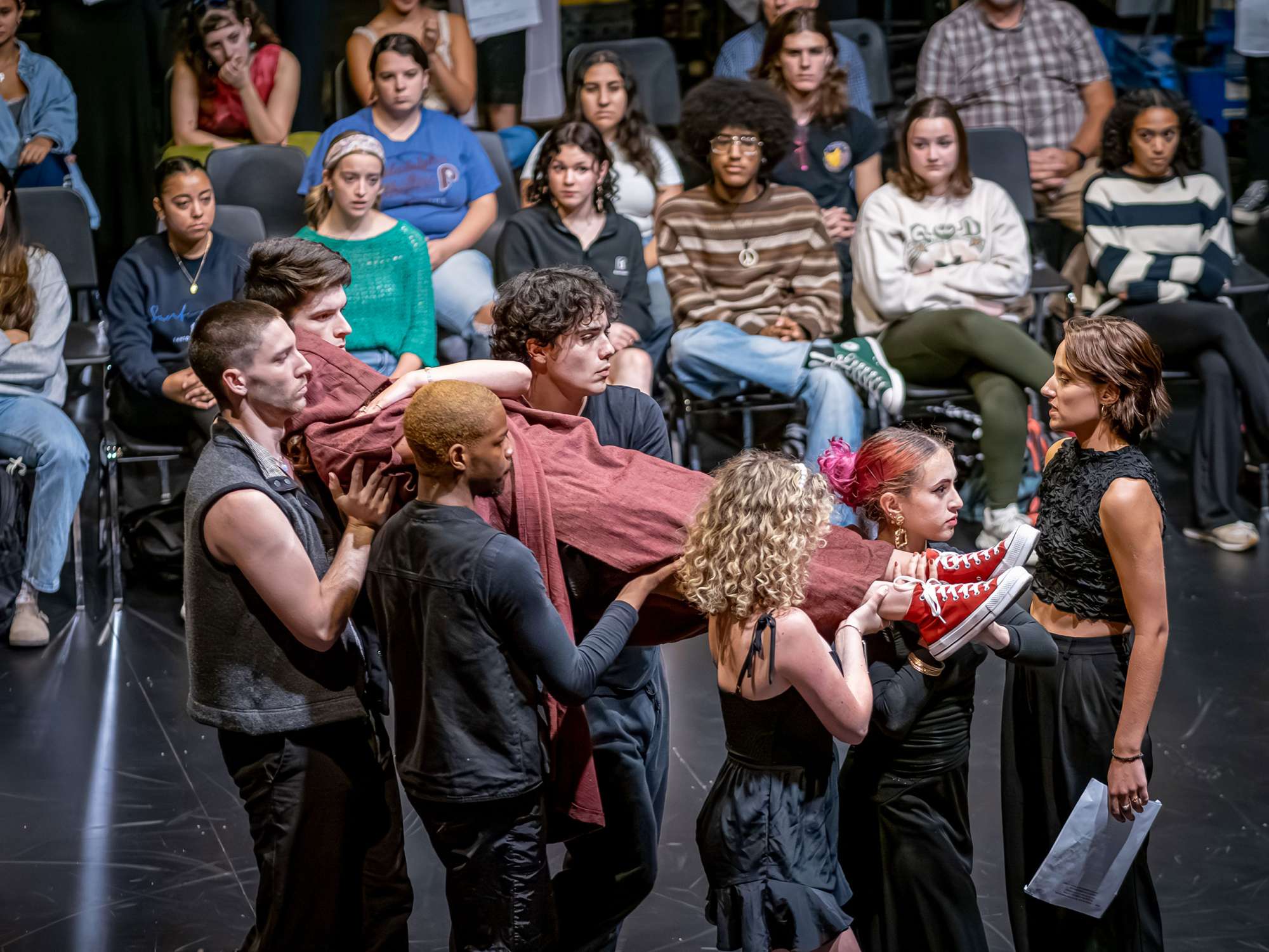 Five actors carry an actor wrapped in cloth as the audience seated on the stage looks on.