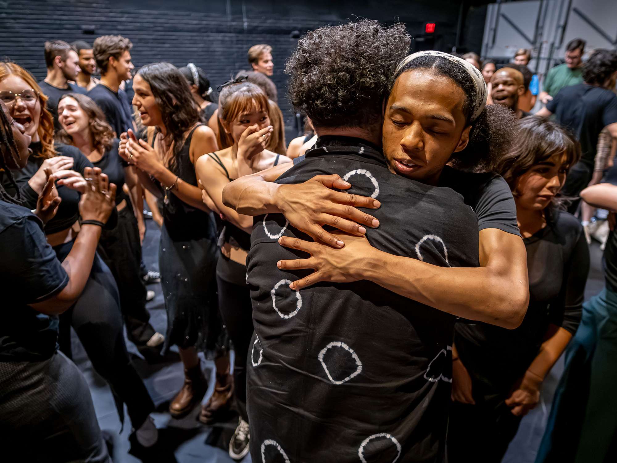 A student actor hugs the director while other student actors gather in the background.