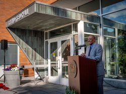 A man in a suit stands at a podium in front of the Irvin D. Reid Hall entrance.
