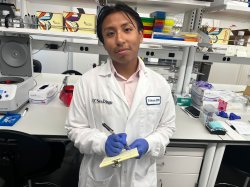 A student wearing a white lab coat writes notes on a small pad of paper inside a science lab.