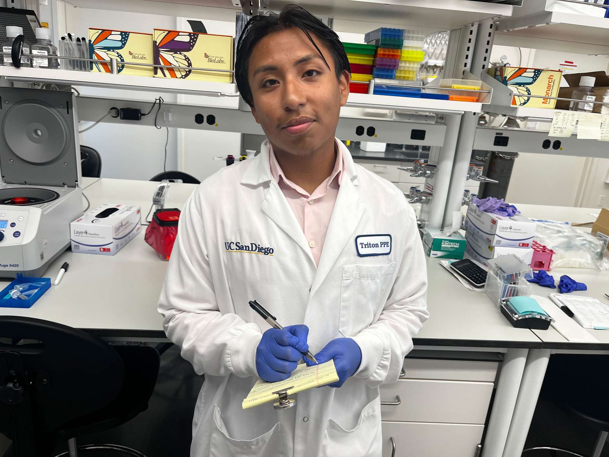 A student wearing a white lab coat writes notes on a small pad of paper inside a science lab.