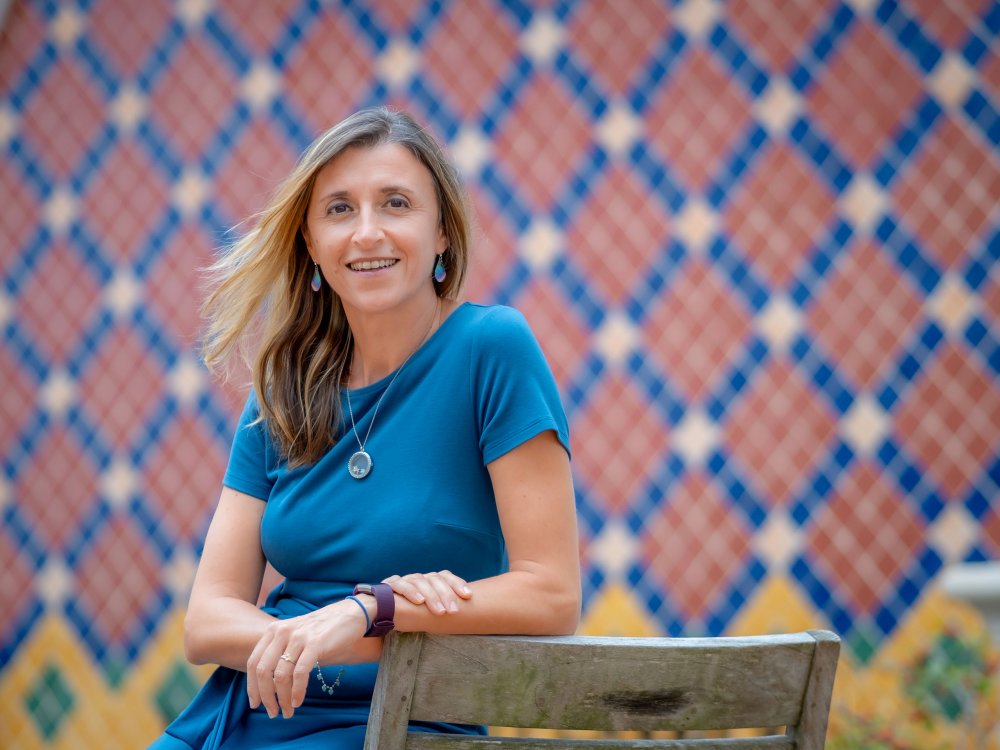 Antonella Calarota-Ninman poses for a portrait on a wooden chair in front of colorful mosaic tiles.