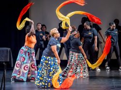 Three women dance with colorful scarves while students dance behind them.