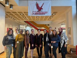 Nine students and a professor stand under a Montclair State University banner with Red Hawk displayed.