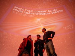 Three people stand on a stage with the words "Space Talk: Cosmic Clues From Gravitational Waves" on the domed ceiling behind them.
