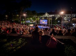 An audience in an outdoor amphitheater watch a movie with a symphony on campus.