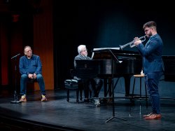 A student plays trumpet while Arturo Sandoval looks on. The trumpet player is accompanied by a pianist.