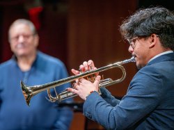 A student plays trumpet for Arturo Sandoval, who smiles in the background.