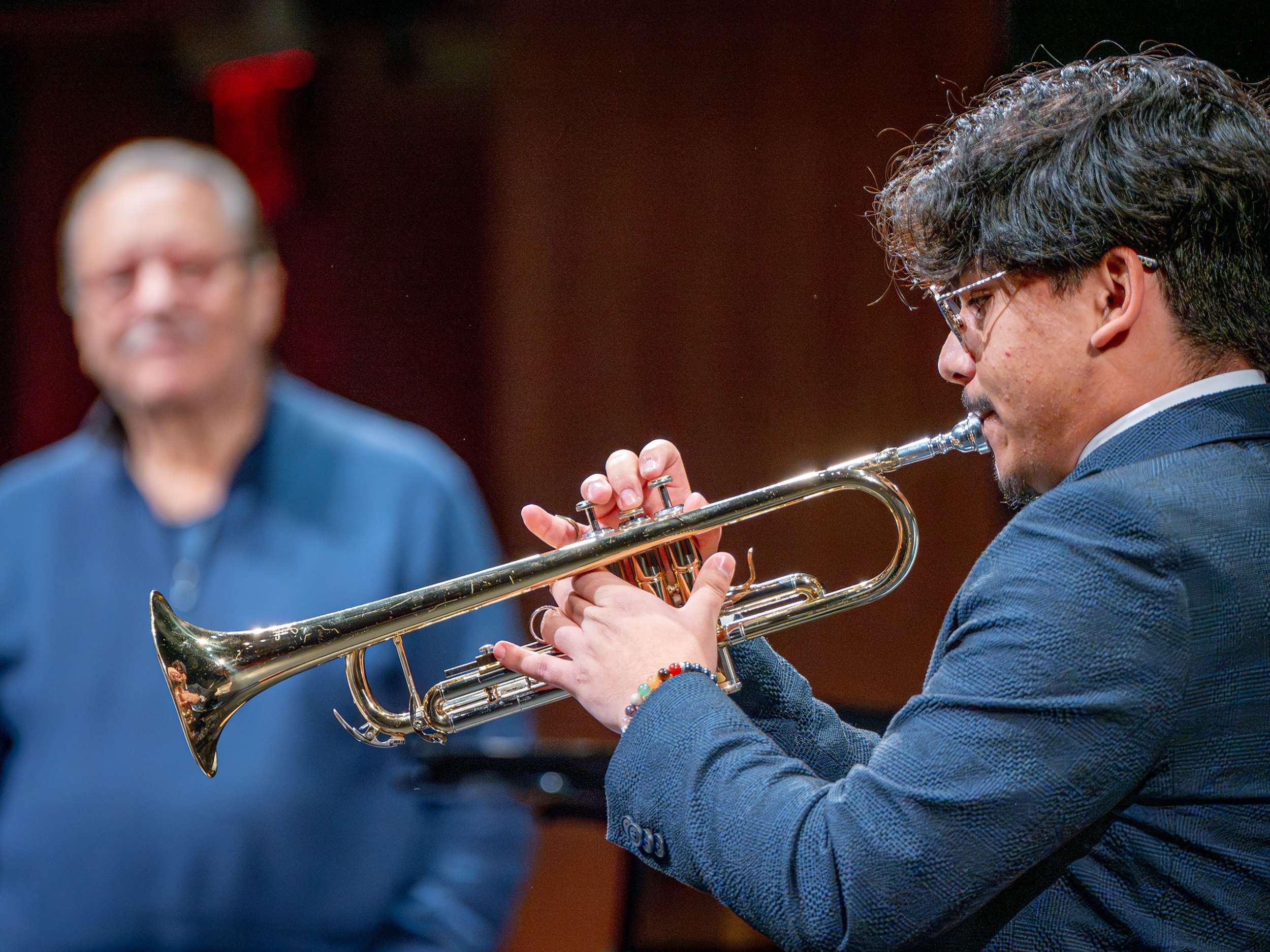 A student plays trumpet for Arturo Sandoval, who smiles in the background.