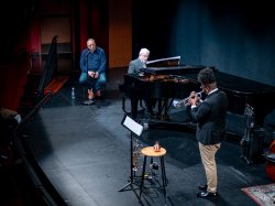 A student plays a trumpet onstage for Arturo Sandoval. A professor accompanies the student on piano.