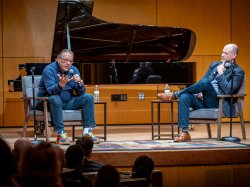 Arturo Sandoval sits in a chair and speaks into a microphone while on stage with Anthony Mazzocchi.