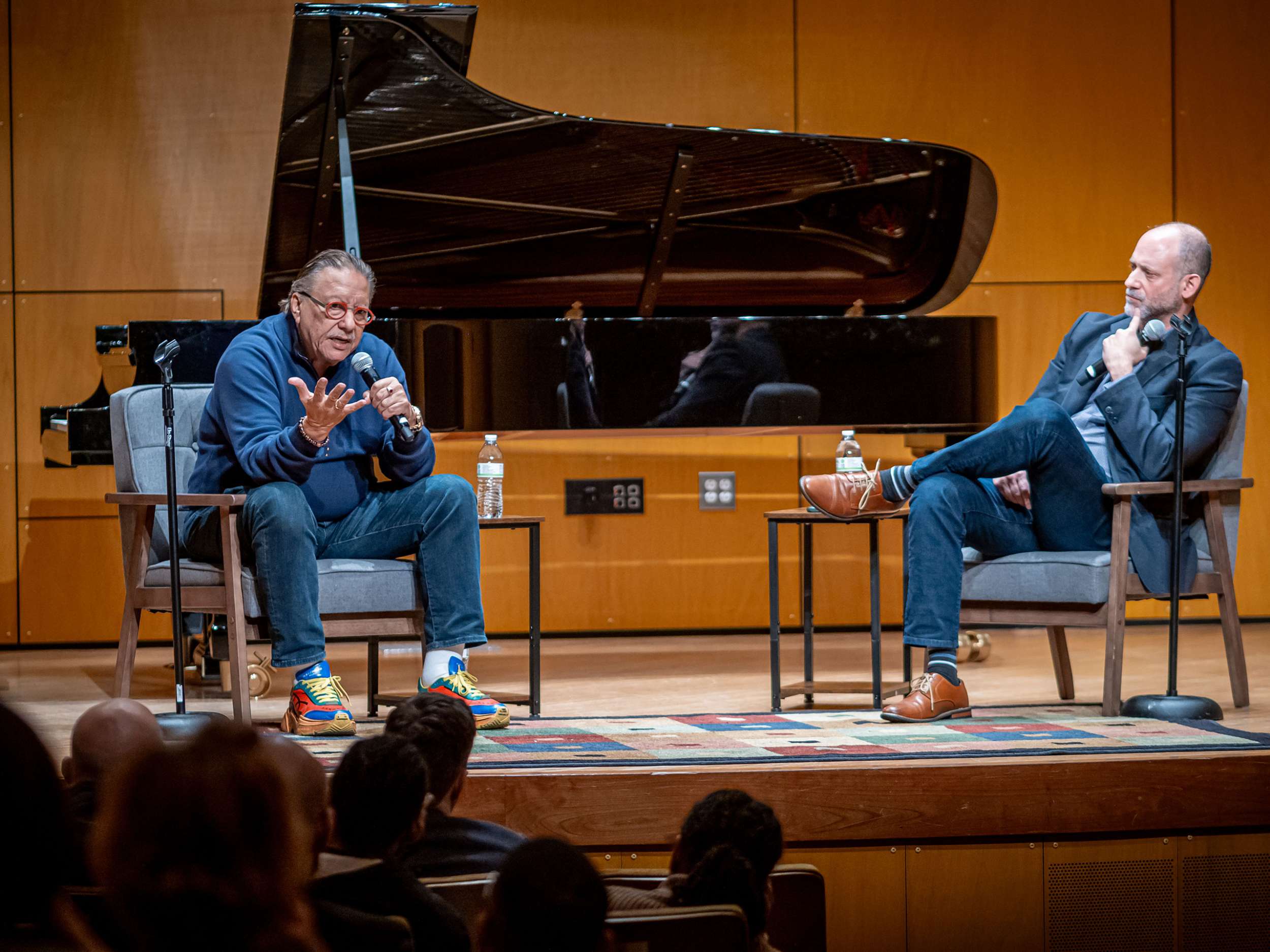 Arturo Sandoval sits in a chair and speaks into a microphone while on stage with Anthony Mazzocchi.