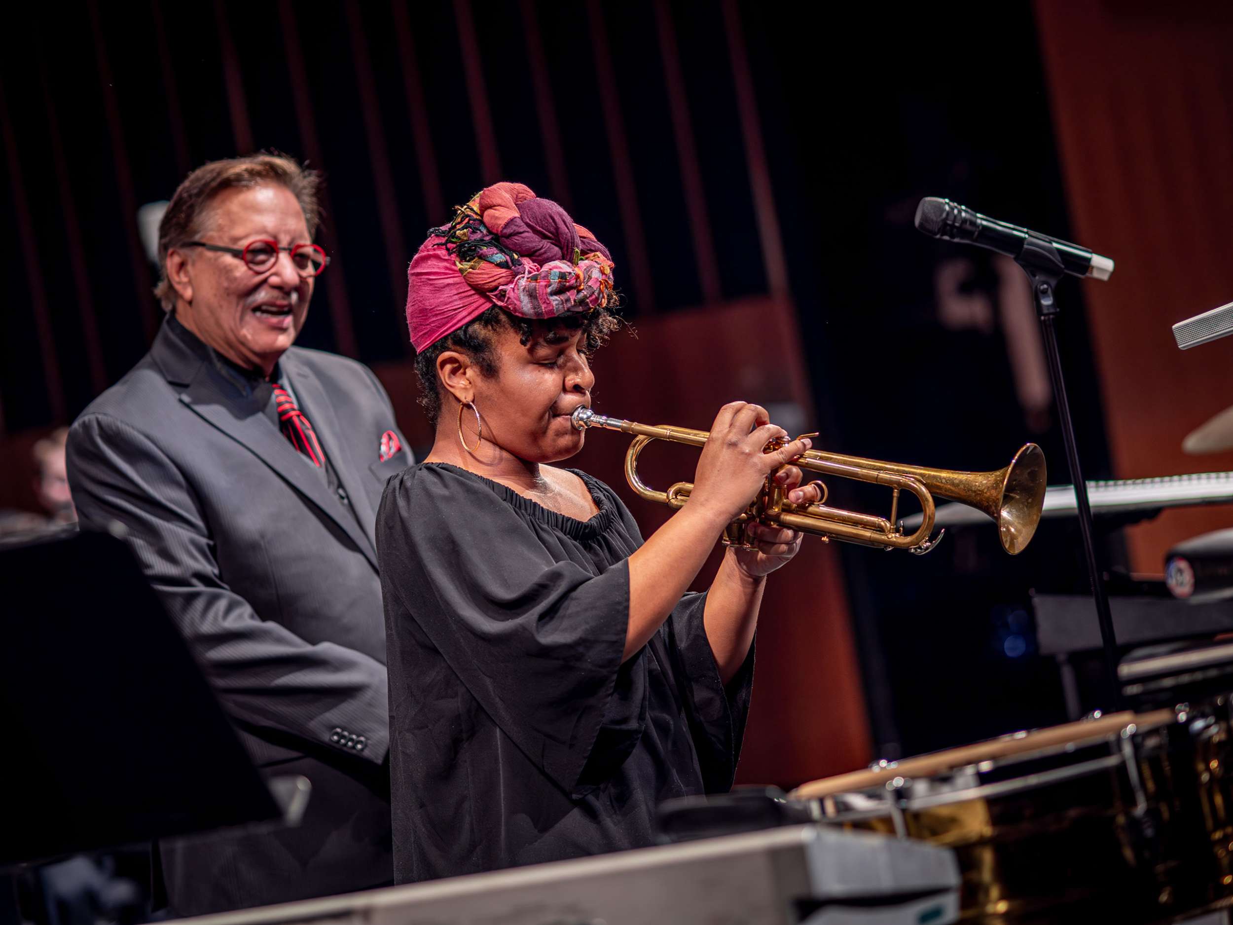 A woman wearing a colorful headscarf plays trumpet as Arturo Sandoval smiles behind her.