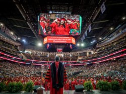 A man in robes addresses an arena audience with graduates projected on a jumbo-tron.