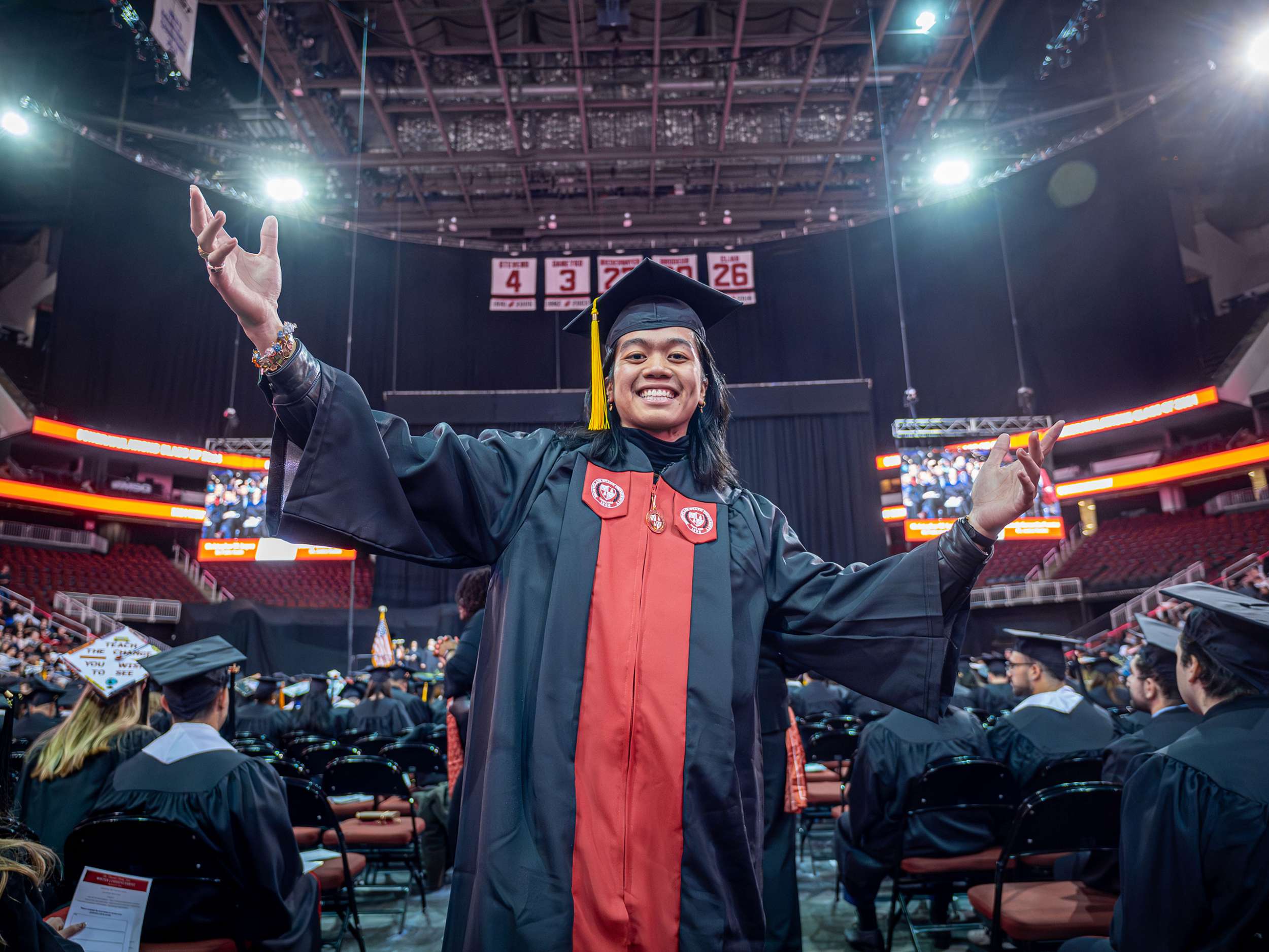 A student in a cap and gown smiles and raises his arms.