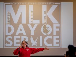 A student with an outstretched arm speaks into a microphone in front of an MLK Day of Service background.