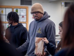 A young man in a sweat shirt that says Bloomfield college passes a sack lunch in an assembly line.