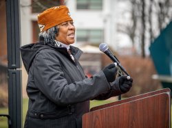 A woman wearing a African head covering, and a coat and gloves speaks into a microphone outdoors.