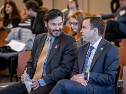 Two men wearing suits chat while seated side by side.