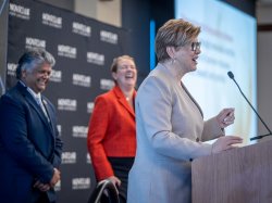 A man and woman laugh while a woman speaks at a lectern.