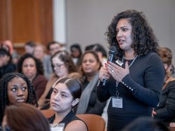 A woman in a black dress stands amid an audience speaking into a microphone.