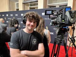 A student stands next to a camera and wears headphones with a red carpet in the background.