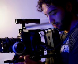 A student looks into a movie camera, bathed in purple light.