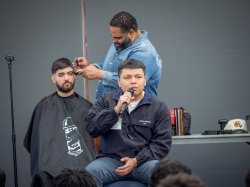 A student speaks into a microphone while another, seated behind him, gets a haircut from a barber.