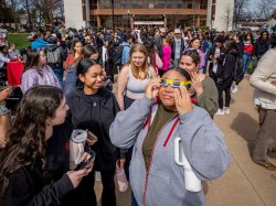 A crowd of students gather, with one looking through solar safety glasses.