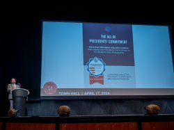 A man at a podium on stage in front of a screen title The All In Presidents' Challenge.
