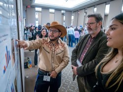 A student wearing a cowboy hat reviews his poster as two people look on.