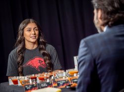 A woman wearing a Red Hawk logo t-shirt, interview President Koppell over chicken wings with spicy sauces.