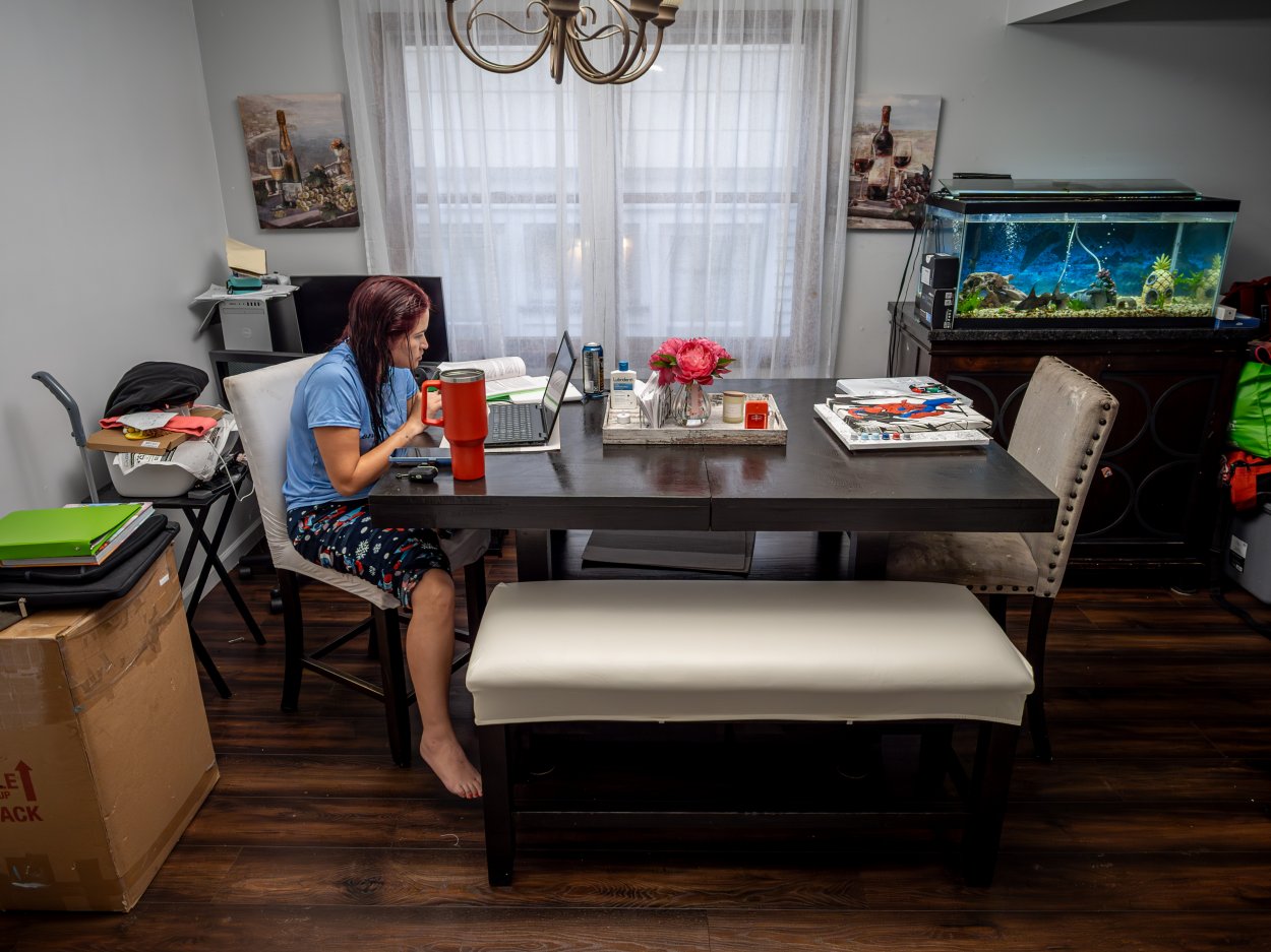 Oscaterin sits in her dining room at sunrise, working on her laptop and writing a discussion post for her class about immigration and families. Study materials, notebooks, and an energy drink are arranged on the table.