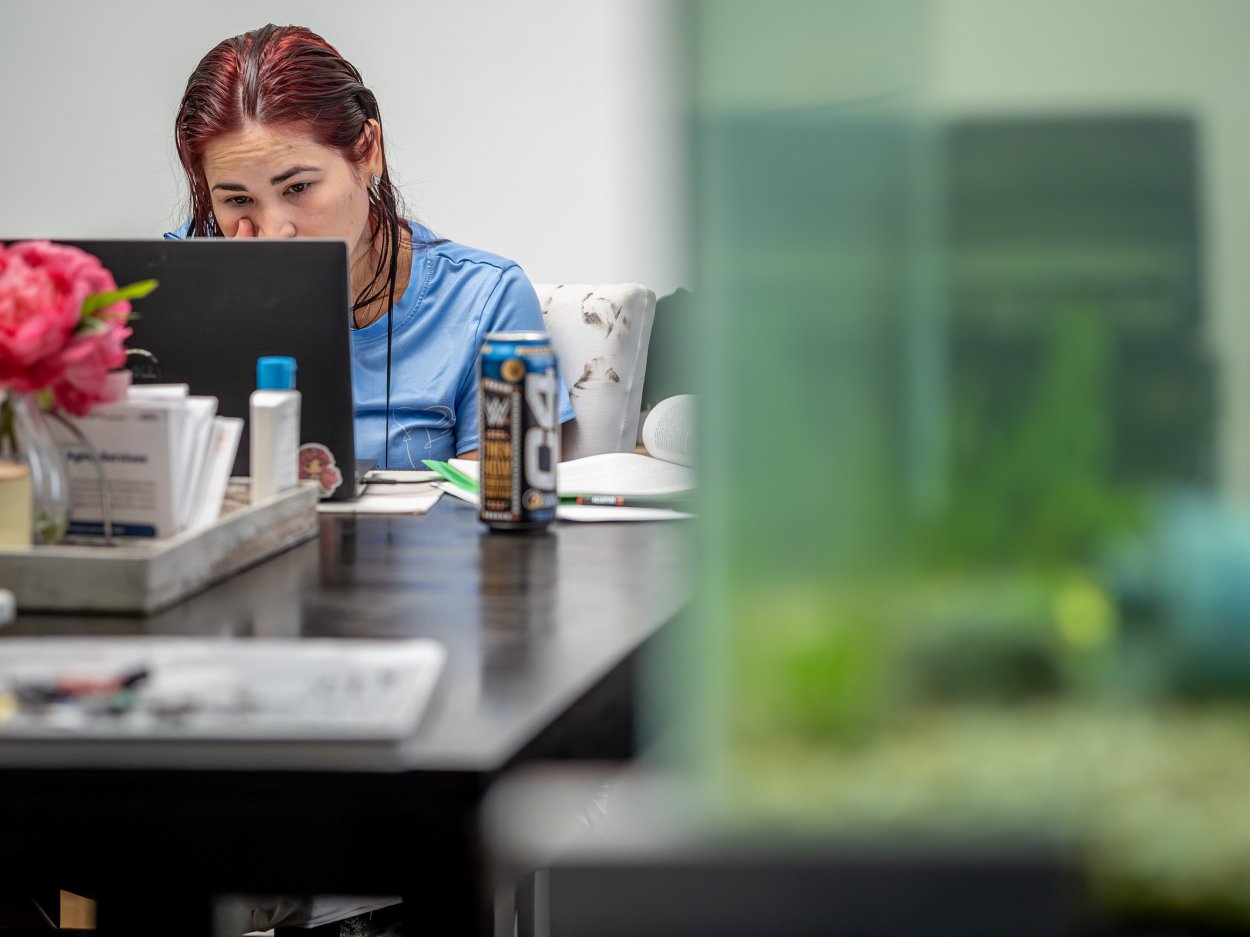 Oscaterin sits at her dining room table, working on her laptop as dawn breaks. An energy drink and study materials are visible on the table as she begins her day focused on her goal of graduating in January 2026.
