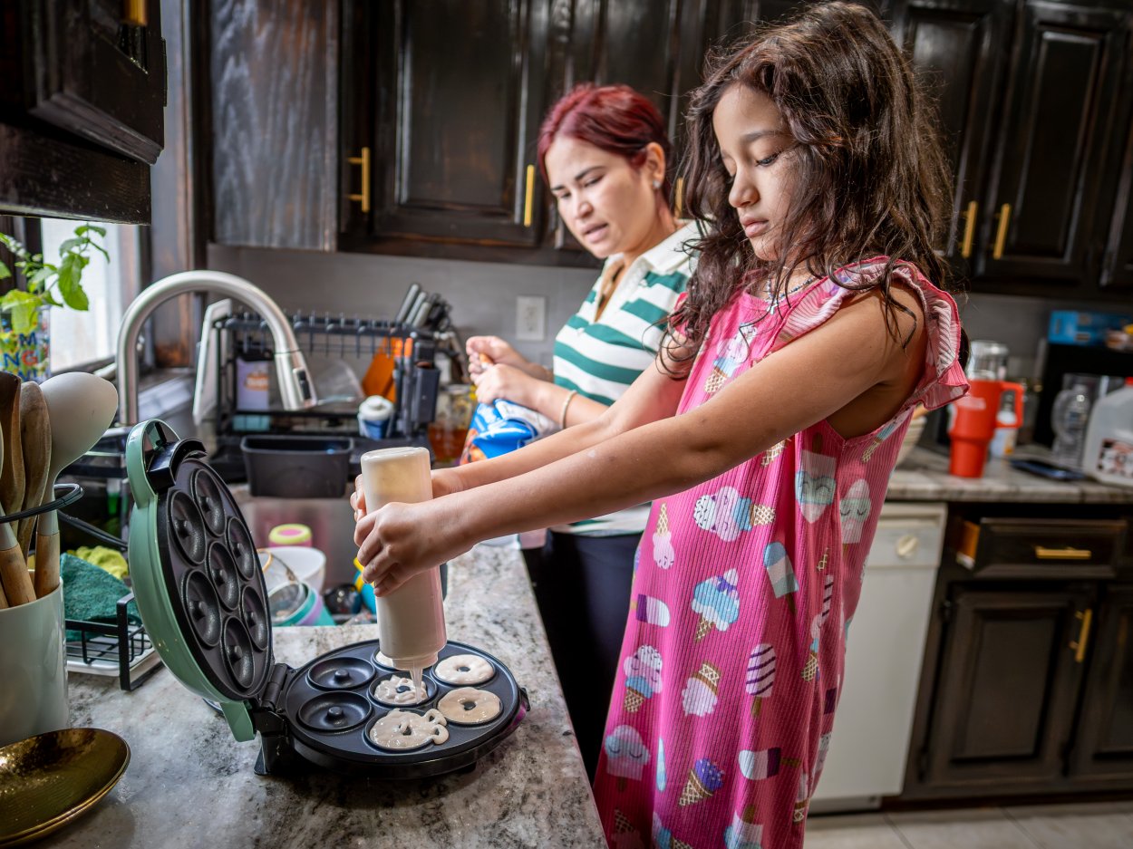 Kaylah helps prepare breakfast in the kitchen, squeezing batter into the donut maker as Oscaterin guides her from behind. Cooking together is part of the family’s morning routine, with everyone pitching in before school and work.