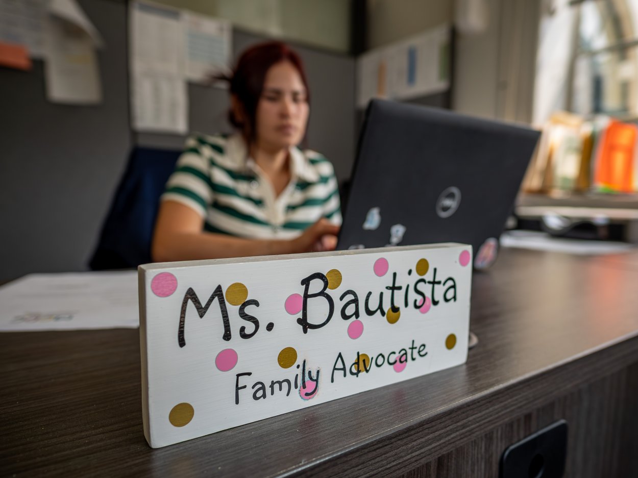 Oscaterin sits at a desk with a laptop, behind a nameplate that reads "Ms. Bautista Family Advocate" at the Montclair Child Development Center.