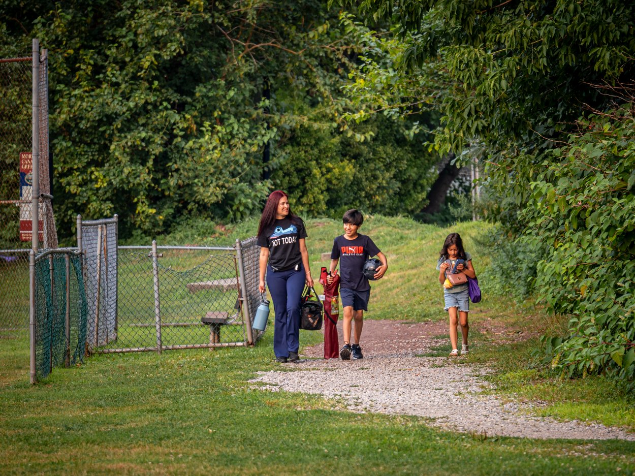 Oscaterin walks along a park path carrying a water bottle, accompanied by two children and sports equipment.