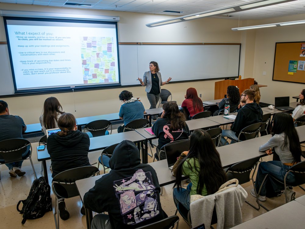 A college classroom with students seated at desks and a professor teaching at the front, a slide projected with class expectations visible on the screen.
