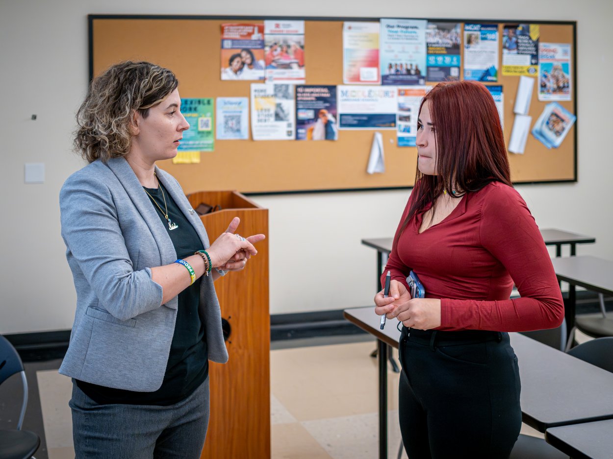 Oscaterin speaks with Assistant Professor Roxanna Ast stand and talk in a college classroom, with a bulletin board and desks in the background.