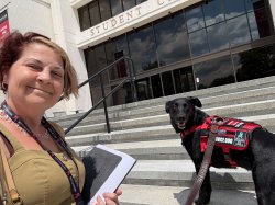 risha Silvasy stands beside Loki, her half Border Collie, half Labrador companion. Loki, a large black dog is wearing a red and gray service harness.