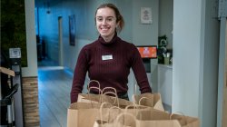 Campus Dietitian and Food Service Manager Jessica Carr, wearing a maroon turtleneck and name badge, stands behind a counter with several brown paper Farm Box bags ready for pickup. The setting is a campus dining facility.
