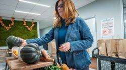 Jennifer Bostedo, wearing a blue jacket and standing indoors, arranges dark green acorn squash and other fresh produce on a wooden table as part of a Farm Box display.