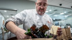 Executive Chef Chris Kelly, dressed in a white chef’s coat, packs a paper bag with fresh ingredients including leafy rainbow chard and a block of cheese. The setting is a campus kitchen.