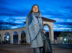 Olga Mikutina bundled in large fringed blue scarf stands outdoors at dusk on a campus walkway.