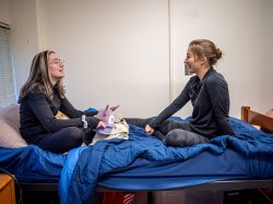 Olga Mikutina and Madison Rowley sit on a bed talking.