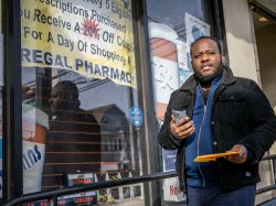 Horatio Wauchope stands outside a neighborhood pharmacy, holding a smartphone in one hand and a yellow envelope in the other, as he walks past a large storefront sign advertising prescription discounts.